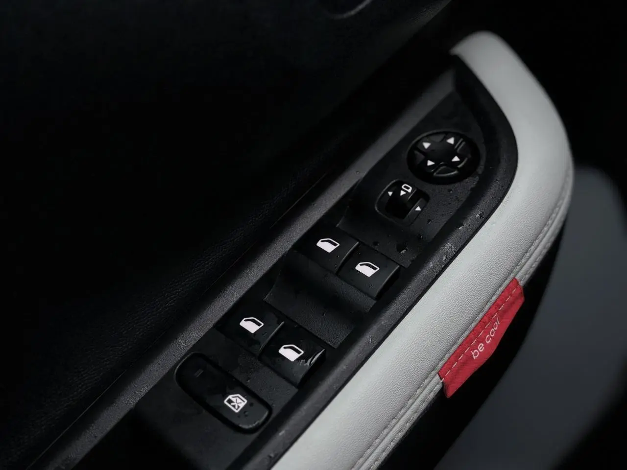 Gros plan sur les commandes des vitres électriques côté conducteur avec surpiqûres blanches et étiquette rouge "be cool" dans l'habitacle.