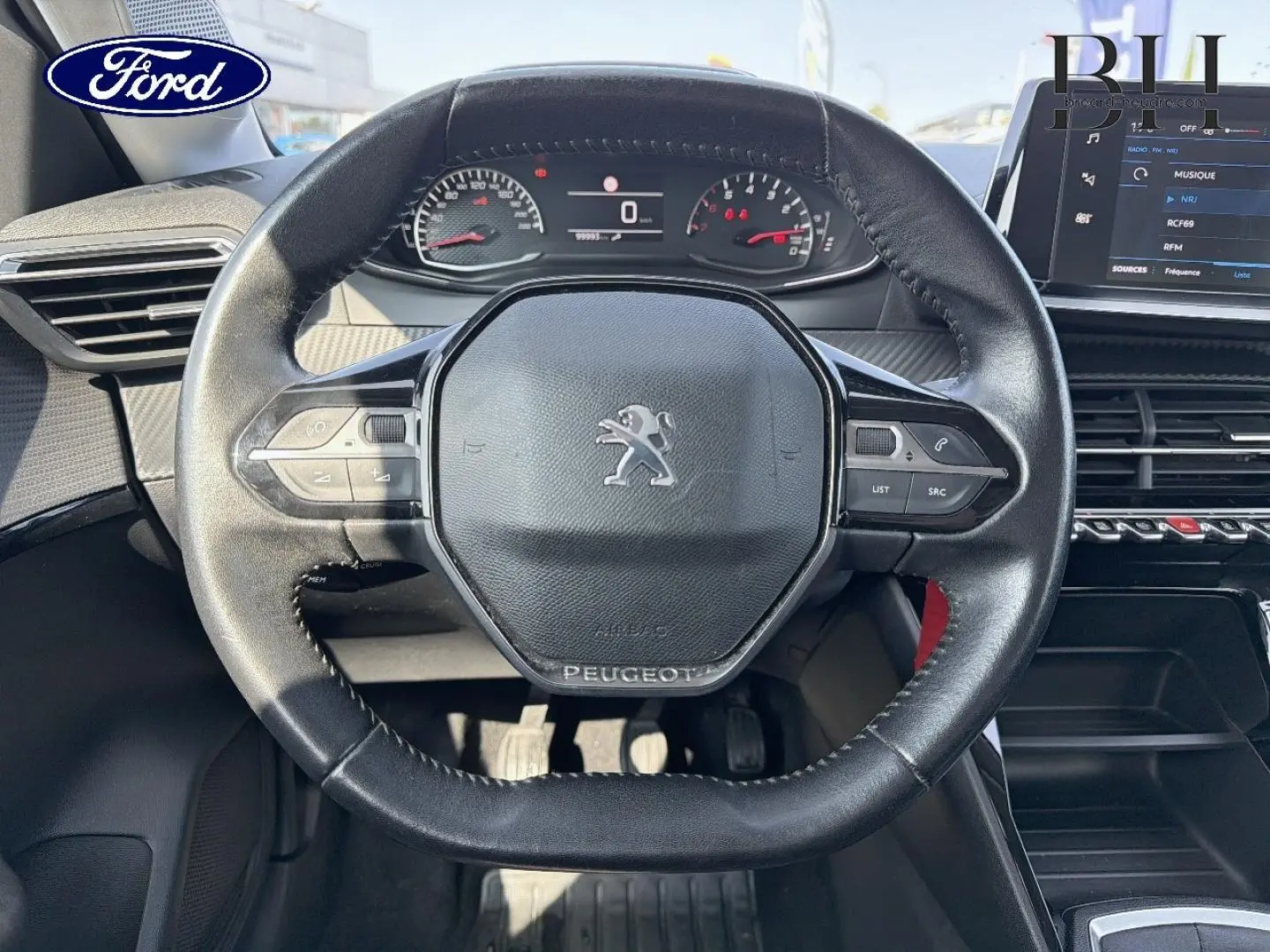 Vue rapprochée du volant noir de la Peugeot 208 1.5 BlueHDi avec tableau de bord et écran tactile visibles.