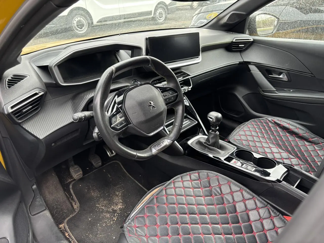Vue intérieure côté conducteur de la Peugeot 208 GT 2021 jaune, avec volant compact, boîte manuelle et sièges noirs matelassés rouges.