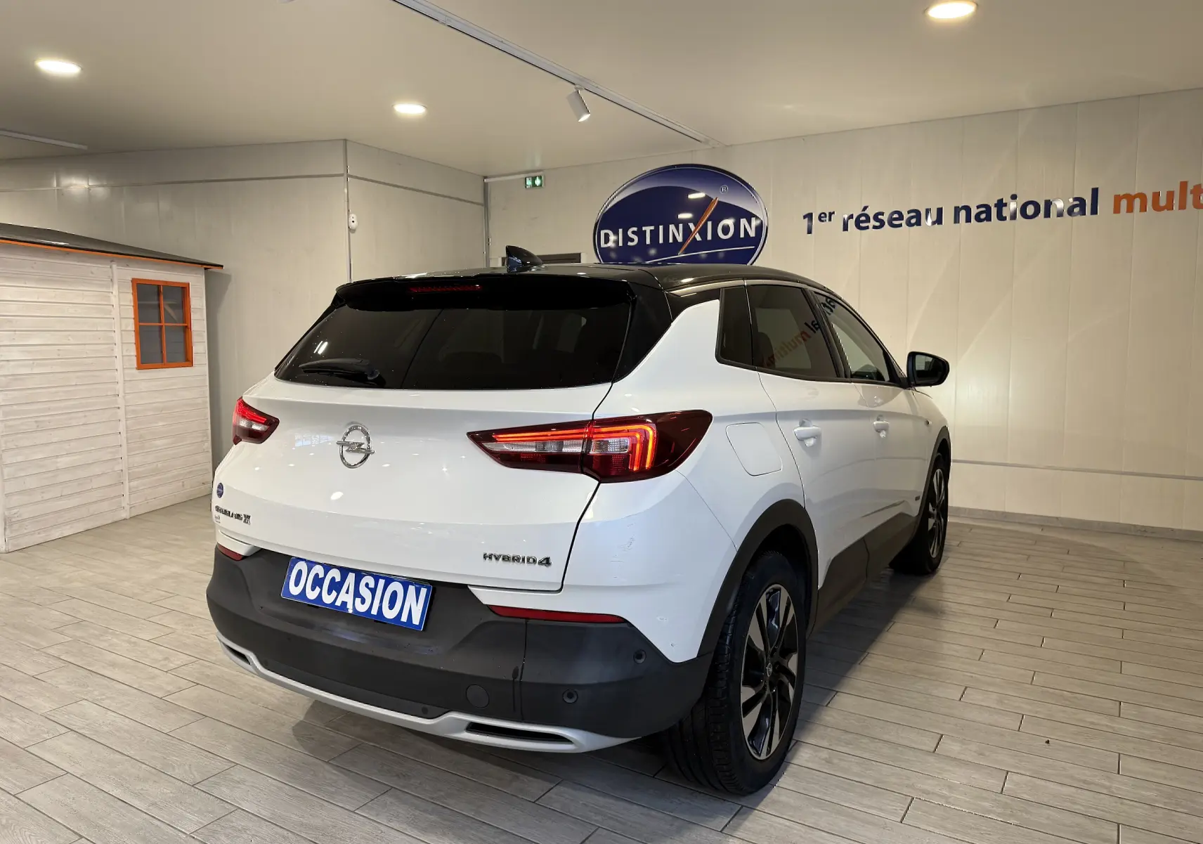 Vue 3/4 arrière droite d’un Opel Grandland X blanc avec toit noir et badge Hybrid4, en intérieur showroom.