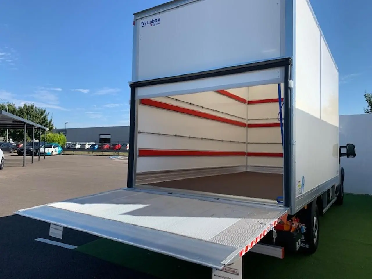 Vue arrière d'un FIAT Ducato blanc avec hayon élévateur déployé et caisse fourgon vide ouverte sur un parking.