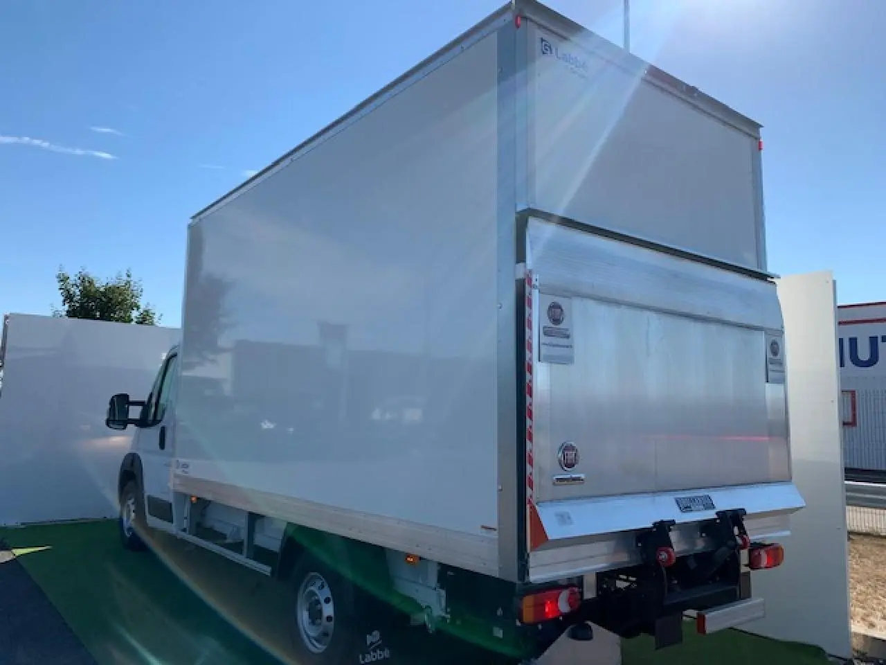 Fiat Ducato Fourgon blanc vu de l'arrière droit, avec hayon élévateur et caisse fermée sous un ciel bleu.