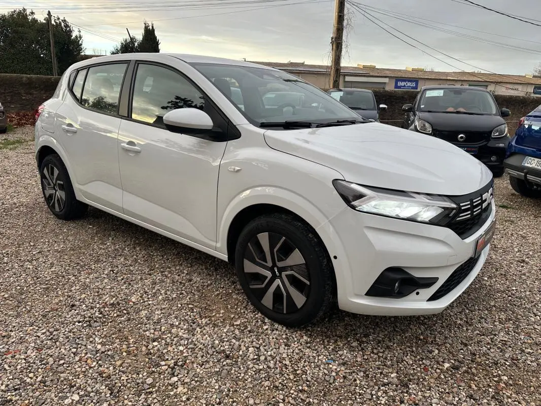 Dacia Sandero blanc vue 3/4 avant droit, avec feux allumés et jantes au design moderne.