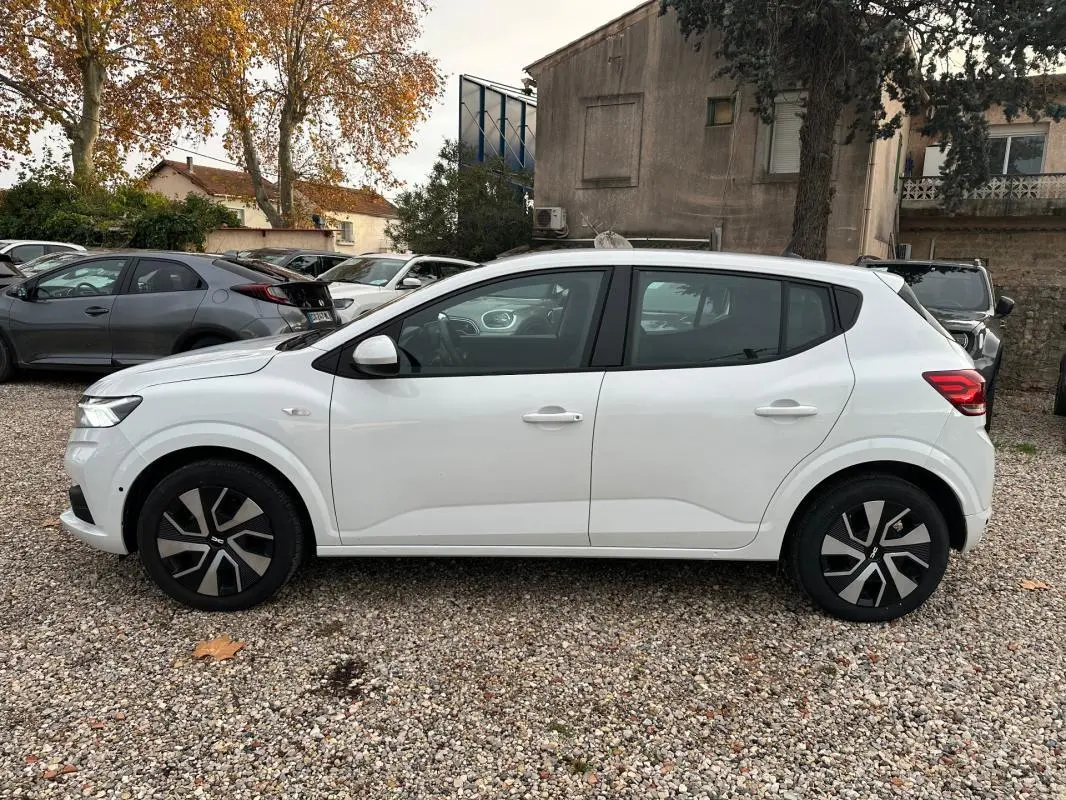 Profil côté gauche d'une Dacia Sandero blanche 2024 garée sur un sol gravillonné avec jantes noires et argentées.