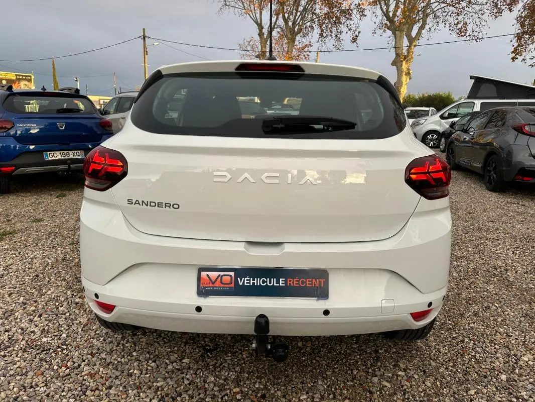 Vue arrière d'une Dacia Sandero blanche 2024 avec feux arrière allumés et attelage de remorque visible.