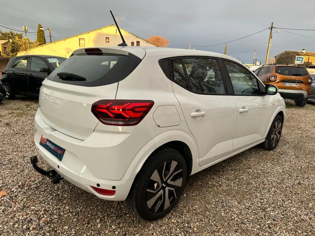 Vue 3/4 arrière droite d'une Dacia Sandero blanche 2024 avec feux arrière allumés et attelage visible.