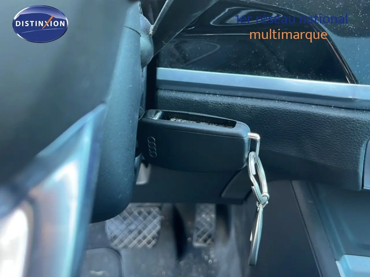 Gros plan sur la clé noire Audi suspendue près du volant, avec les pédales et la console noire en arrière-plan.