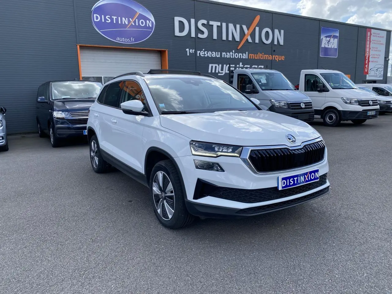 Vue 3/4 avant d'un Skoda Karoq blanc moon métallisé avec jantes alliage, stationné devant un concessionnaire Distinxion.