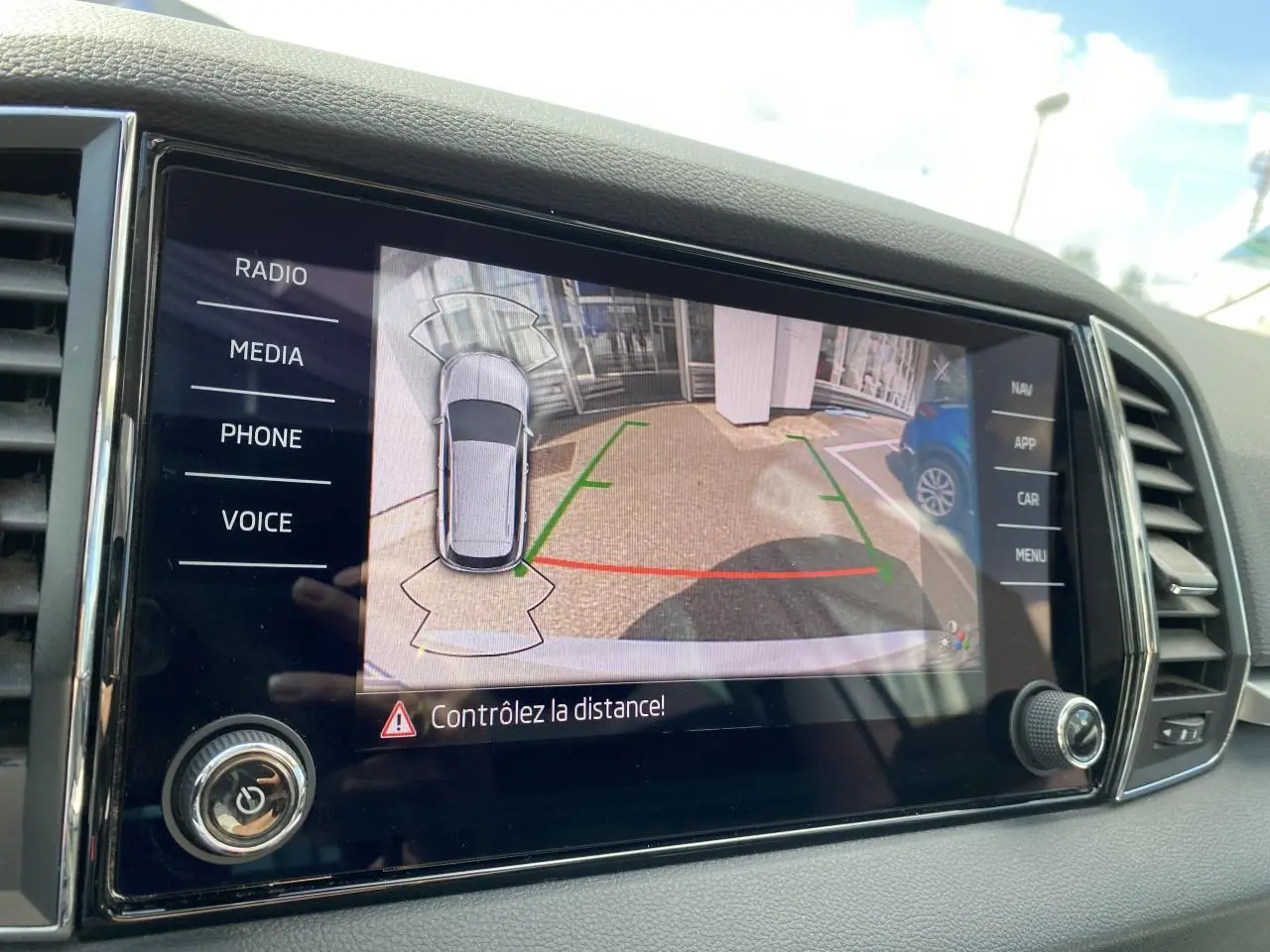 Écran tactile intérieur montrant la caméra de recul avec vue aérienne du Skoda Karoq blanc Moon métallisé en stationnement.