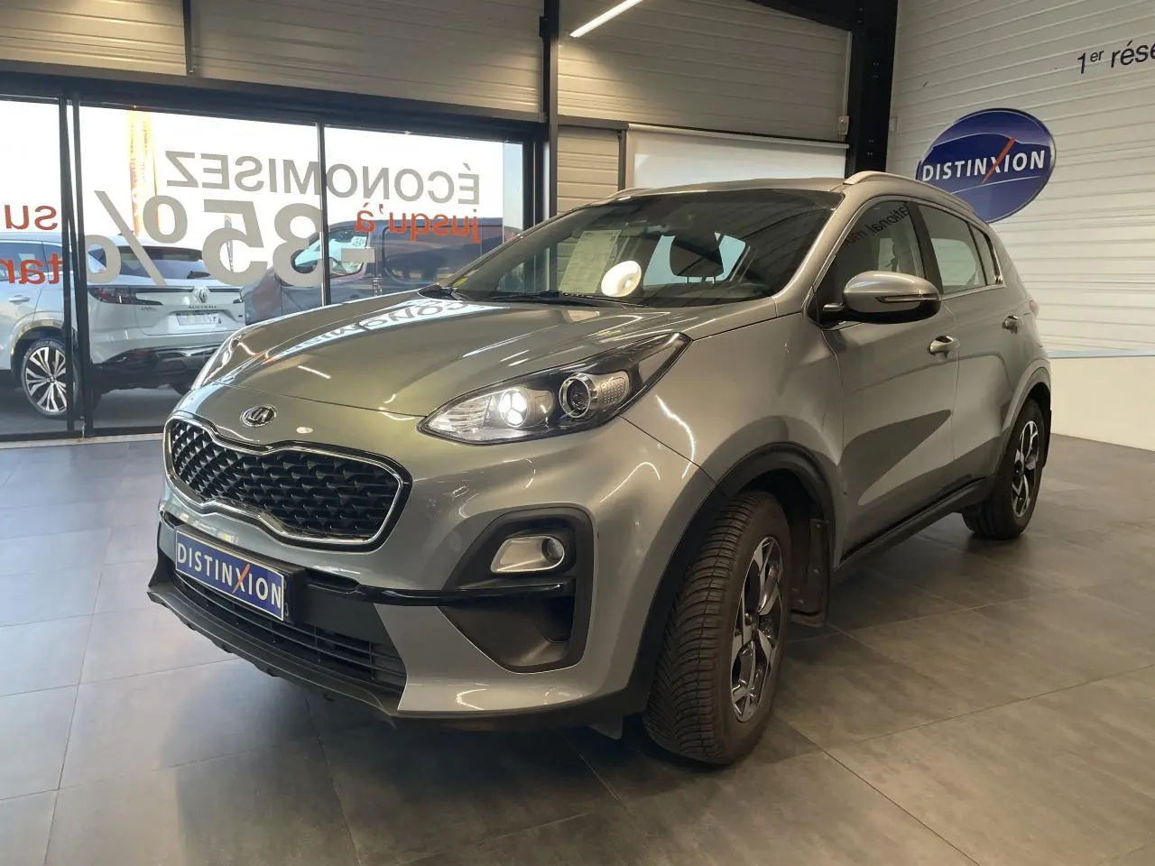 KIA Sportage gris clair en 3/4 avant droit, avec calandre noire mat et jantes alliage 16 pouces visibles.