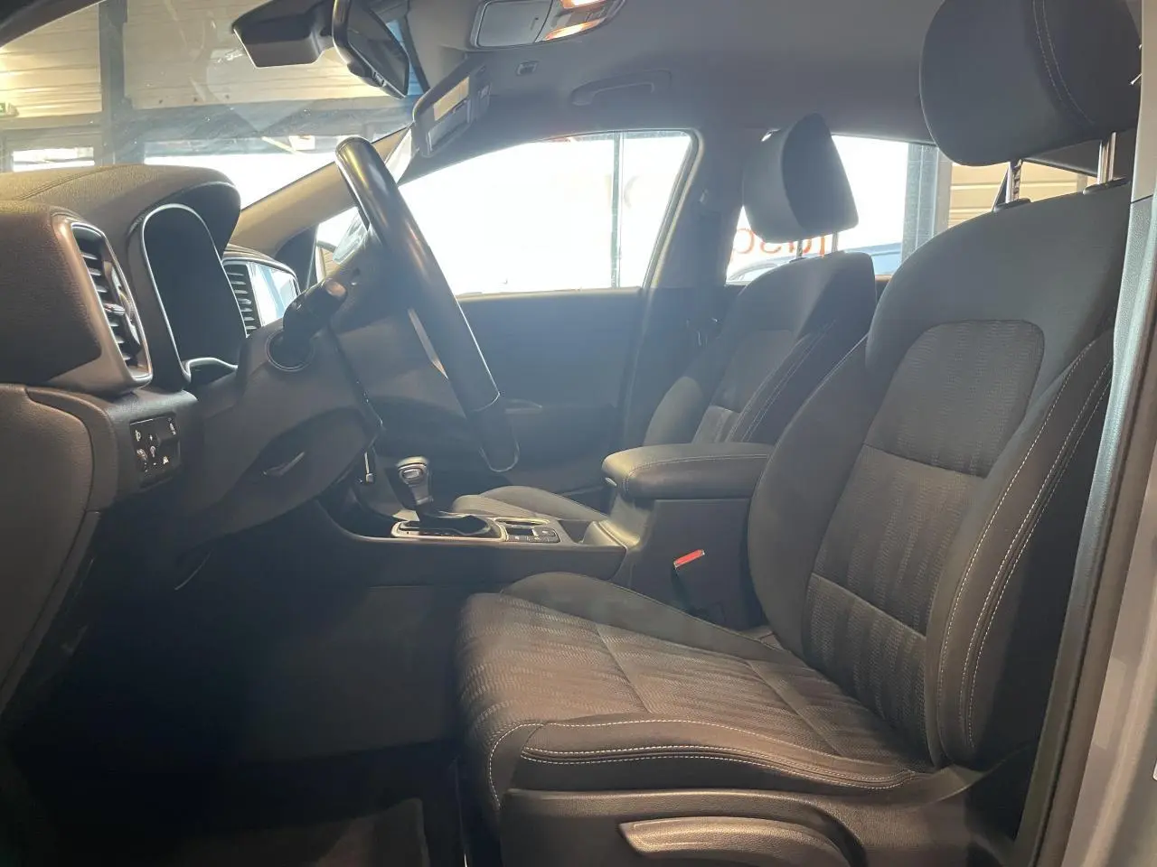 Vue intérieure côté conducteur du Kia Sportage gris clair 2021, mettant en valeur les sièges tissu noirs et l'accoudoir central.