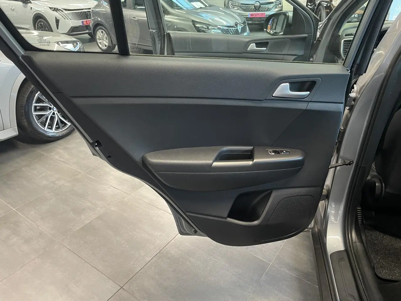 Porte arrière droite ouverte d’un KIA Sportage gris clair, montrant la garniture intérieure noire avec commandes de lève-vitres.