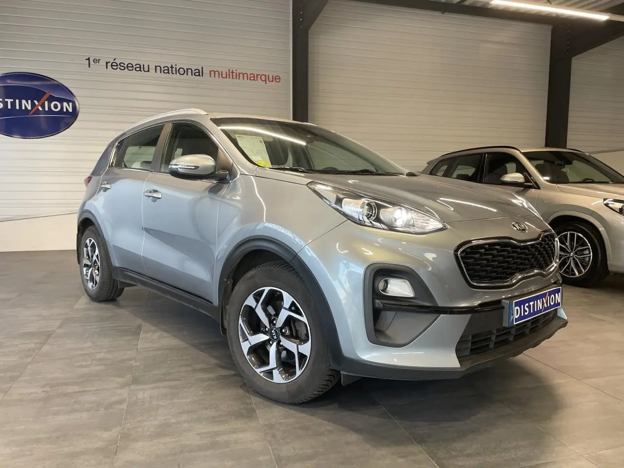 KIA Sportage gris clair en 3/4 avant droit, avec jantes alliage 16 pouces et calandre noir mat.