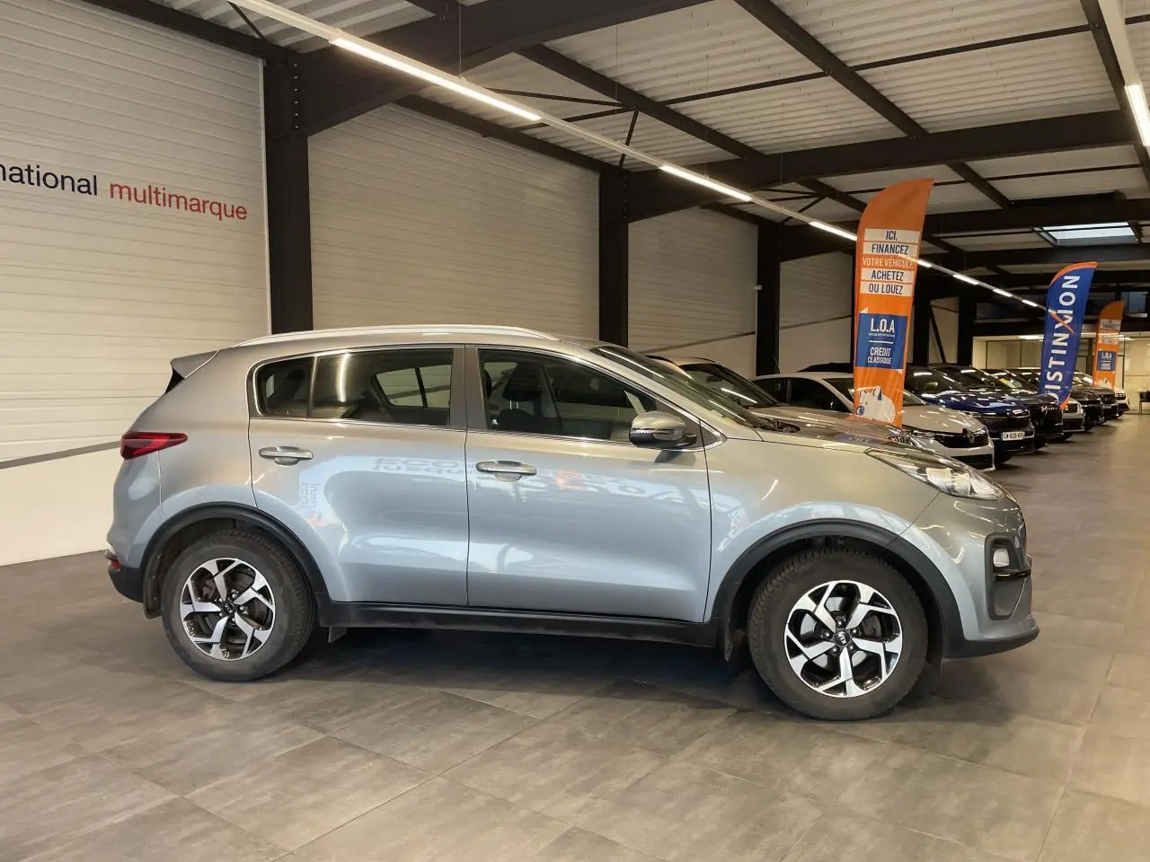 KIA Sportage gris clair vu de profil côté gauche, avec jantes alliage 16 pouces et poignées couleur carrosserie.
