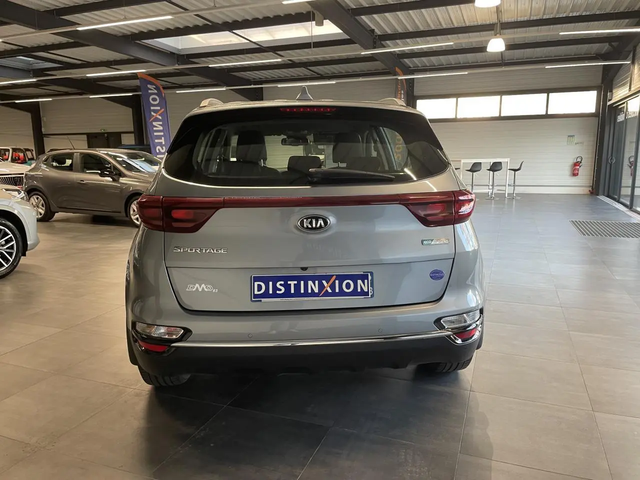 Vue arrière d'un KIA Sportage gris clair en showroom, avec feux arrière reliés par une bande rouge et logo central.