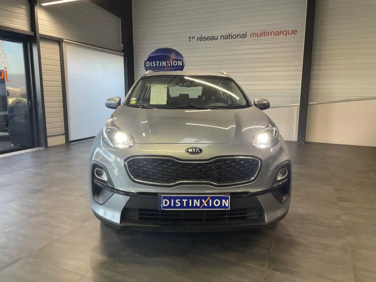 Vue avant d'un KIA Sportage gris clair avec phares allumés, calandre noire et plaque Distinxion en intérieur showroom.