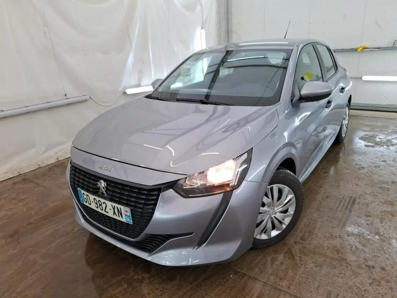 Peugeot 208 gris Artense vue 3/4 avant droit avec phares allumés dans un garage fermé.