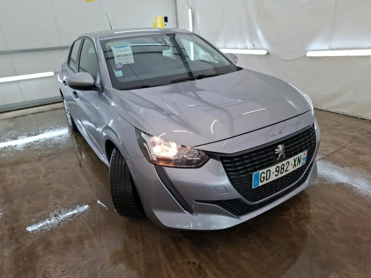 Peugeot 208 gris Artense vue 3/4 avant gauche avec calandre noire et feu avant allumé dans un garage.