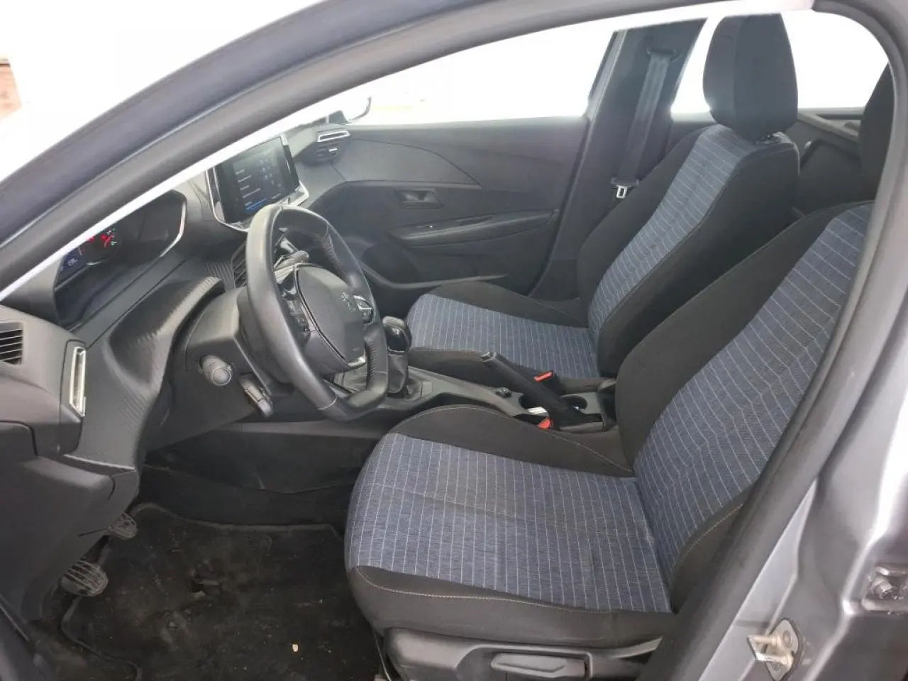 Vue intérieure côté conducteur de la Peugeot 208 gris Artense, montrant les sièges tissu bleu et la planche de bord noire.