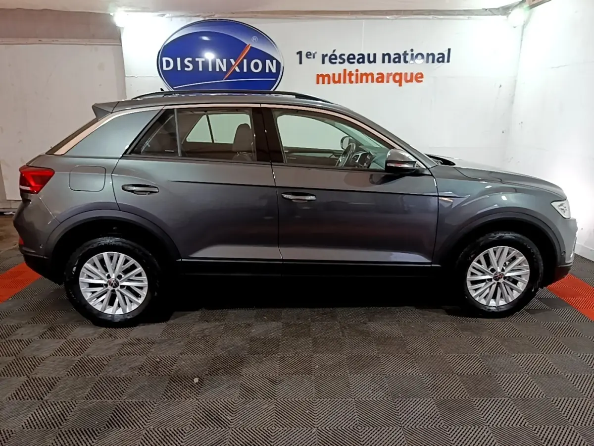 Vue de profil côté gauche d'un Volkswagen T-Roc gris foncé 2022 en intérieur avec jantes alliage et barres de toit noires.