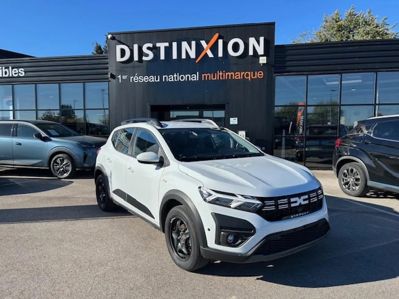 Dacia Sandero Stepway blanc arctique vue 3/4 avant avec calandre noire et barres de toit noires devant un showroom.