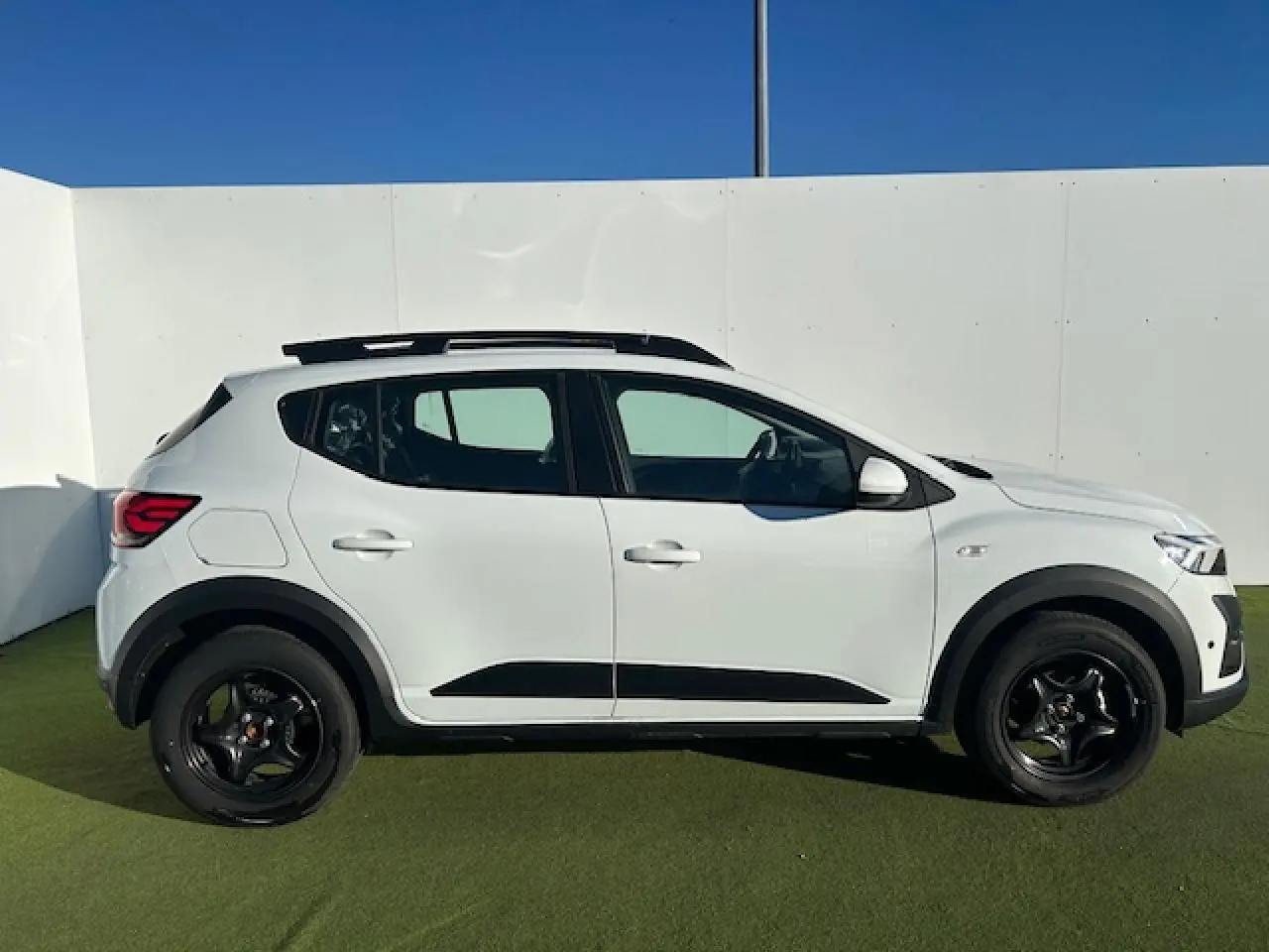 Vue de profil droit d'une Dacia Sandero Stepway blanche avec barres de toit noires et jantes foncées sur fond extérieur.