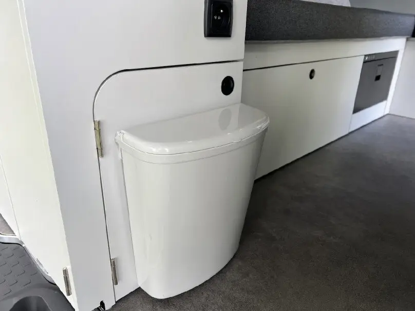 Détail intérieur du fourgon Renault Trafic aménagé, montrant une poubelle blanche intégrée près d’un meuble gris clair.
