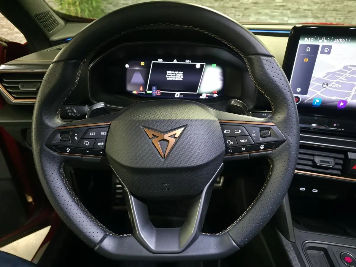 Vue rapprochée du volant en cuir perforé noir avec logo CUPRA cuivre et tableau de bord digital du Formentor rouge 2024.