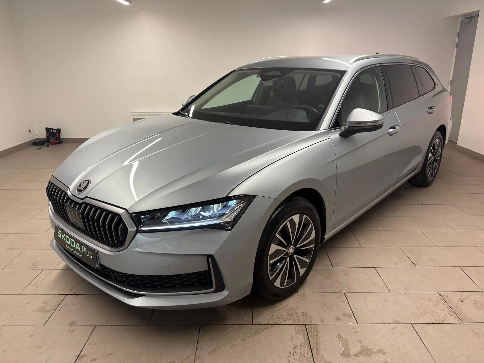Skoda Superb Combi gris argent en 3/4 avant droit, avec calandre noire et jantes alliage distinctives.
