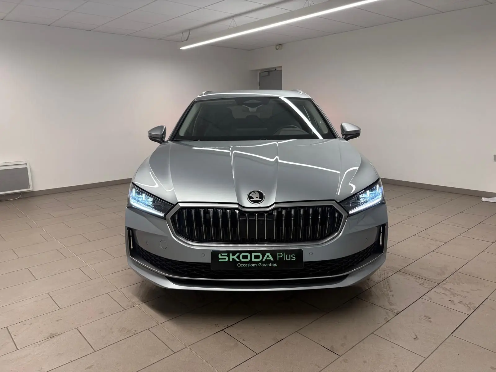 Vue frontale d'une Skoda Superb Combi gris argent 2025 avec calandre noire et phares LED allumés en intérieur.