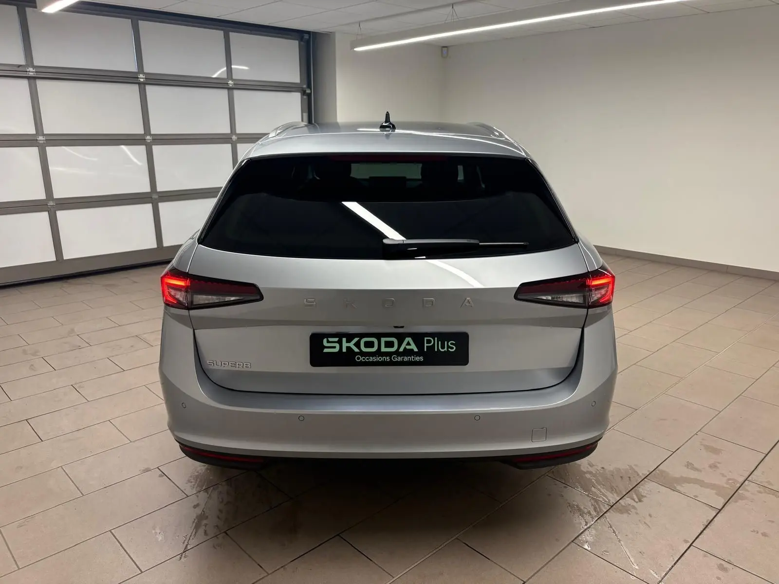 Vue arrière d'un Skoda Superb Combi gris argent 2025 dans un garage, avec feux arrière allumés et logo SKODA visible.