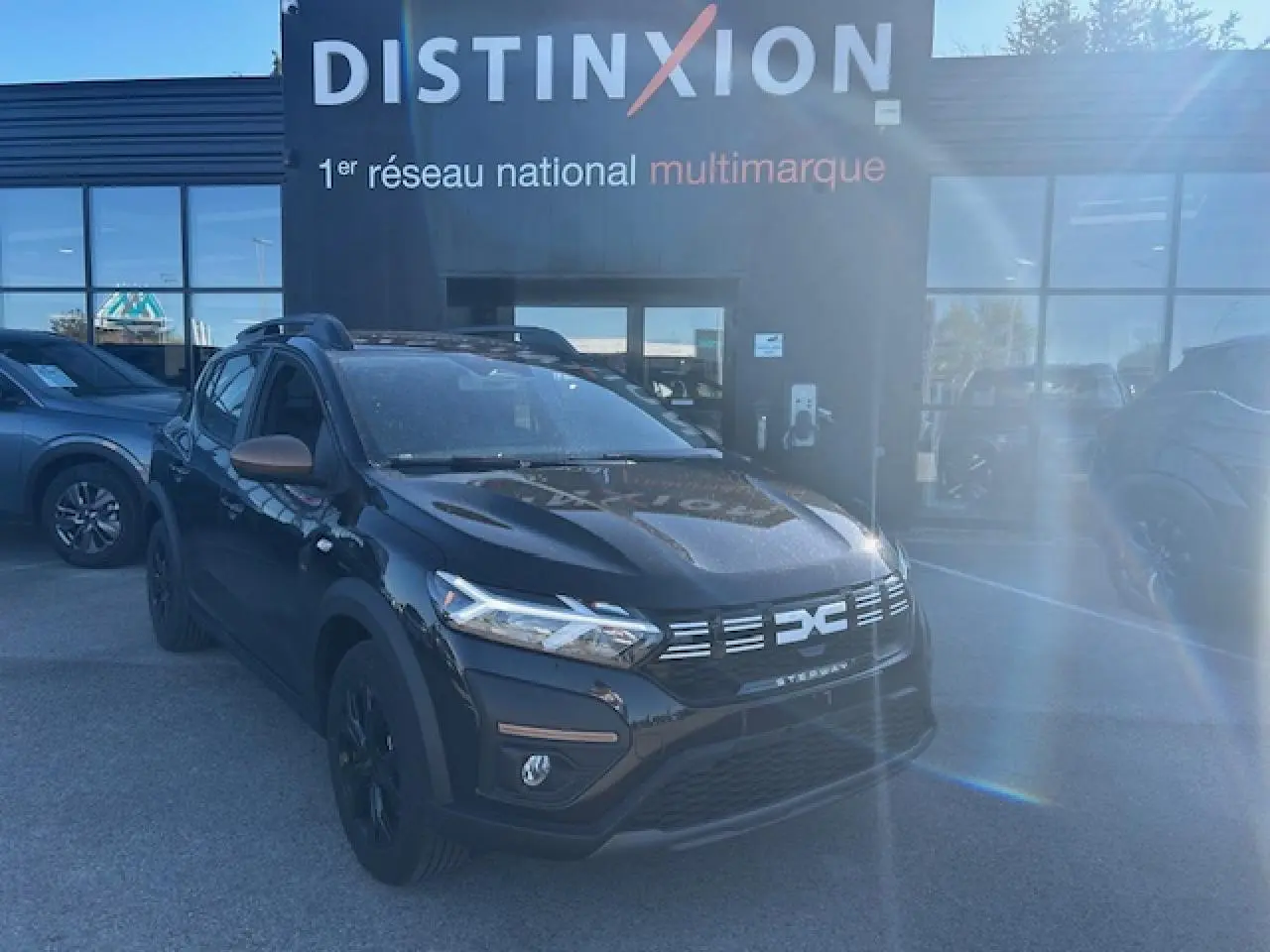 Vue 3/4 avant d'une Dacia Sandero Stepway noire nacrée avec calandre et rétroviseurs brun cuivré, stationnée devant un bâtiment.