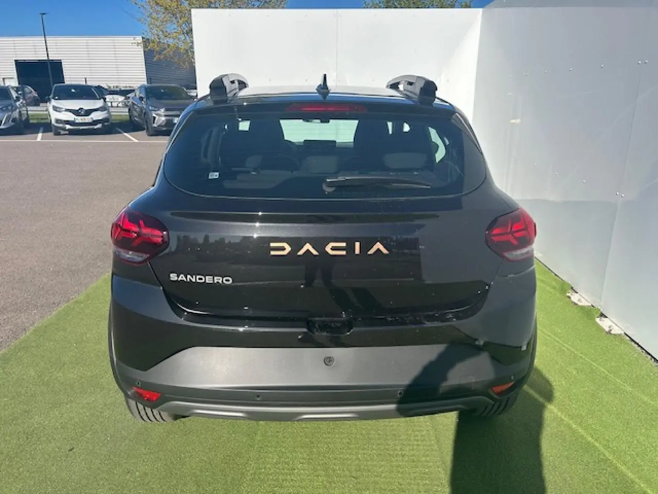 Vue arrière d'une Dacia Sandero III Stepway noir nacré avec logo Dacia brun cuivré et barres de toit visibles.