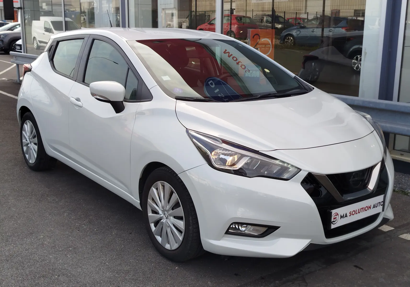Nissan Micra blanche vue 3/4 avant droit, avec phares allumés et jantes alliage, stationnée devant un showroom.