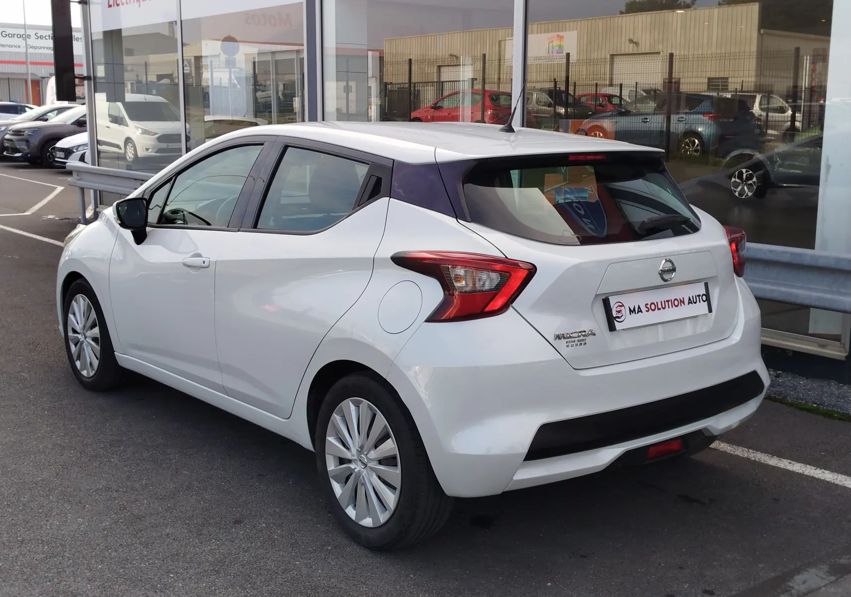 Vue 3/4 arrière droite d'une Nissan Micra blanche 2019 garée devant un showroom avec feux arrière distinctifs.