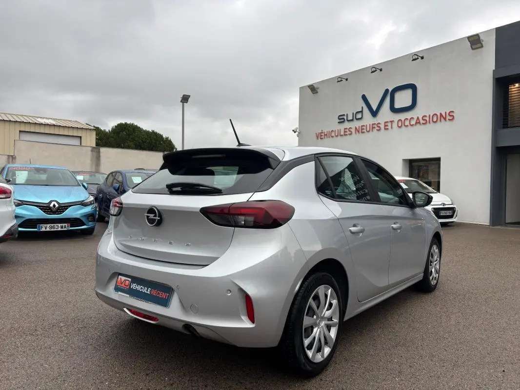 Opel Corsa gris clair vue 3/4 arrière droit, avec jantes classiques et feux arrière foncés, stationnée devant un concessionnaire.