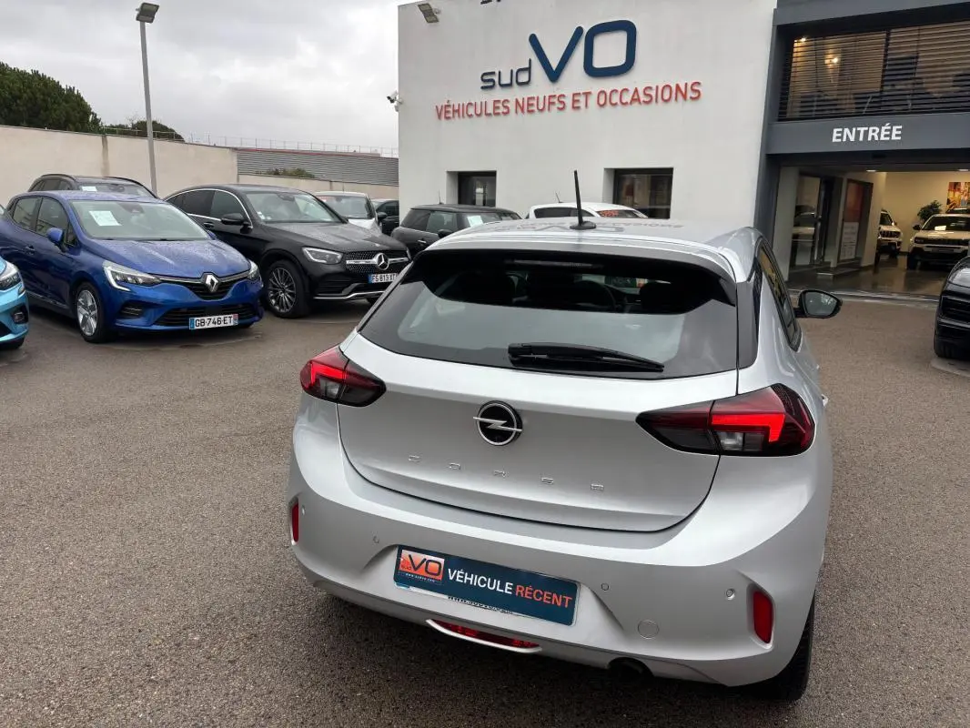 Vue 3/4 arrière d’une Opel Corsa gris clair 2024 garée devant un showroom avec feux arrière allumés.