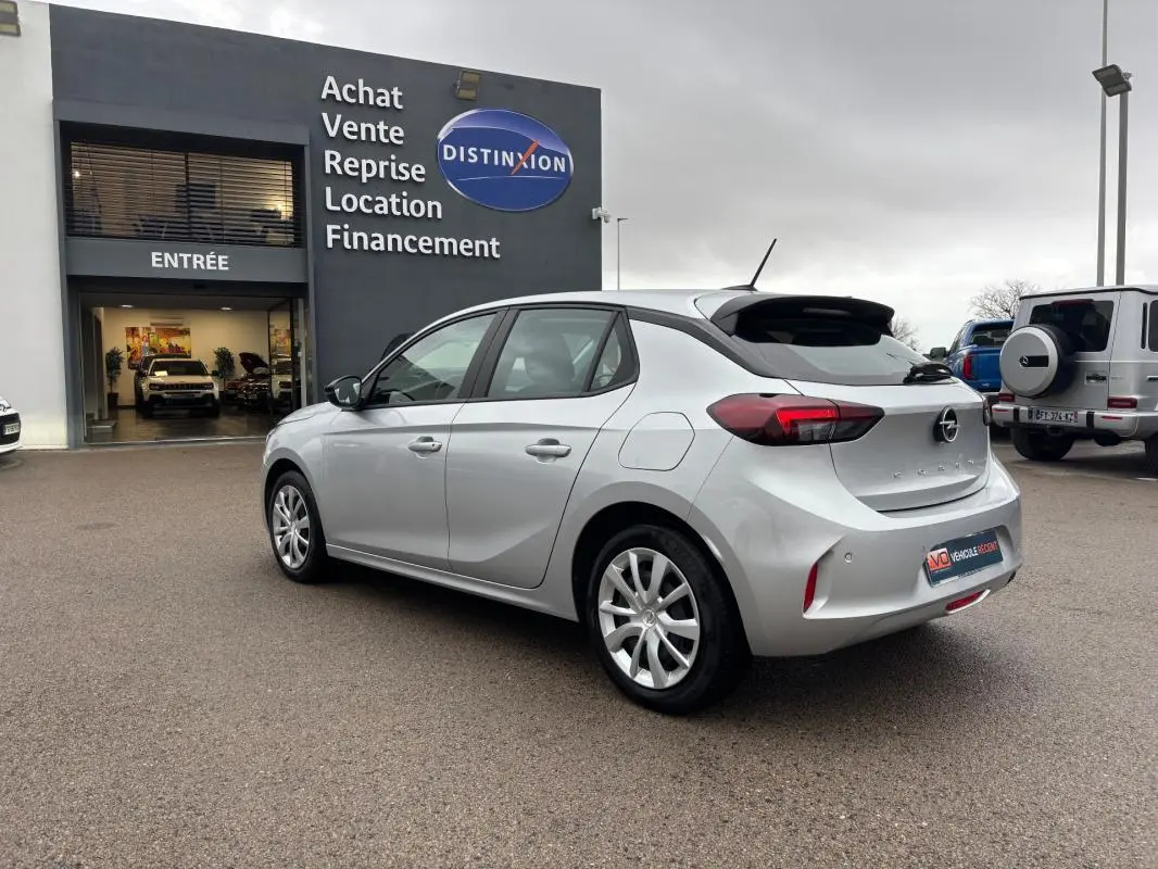 Opel Corsa gris clair vue 3/4 arrière droit, avec jantes classiques 16 pouces, stationnée devant un garage.