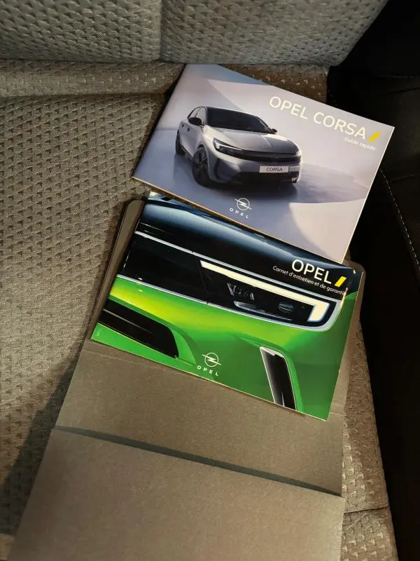 Deux brochures Opel Corsa posées sur un siège en tissu gris, l'une montrant une Corsa grise de face, l'autre un gros plan sur un phare avant vert.