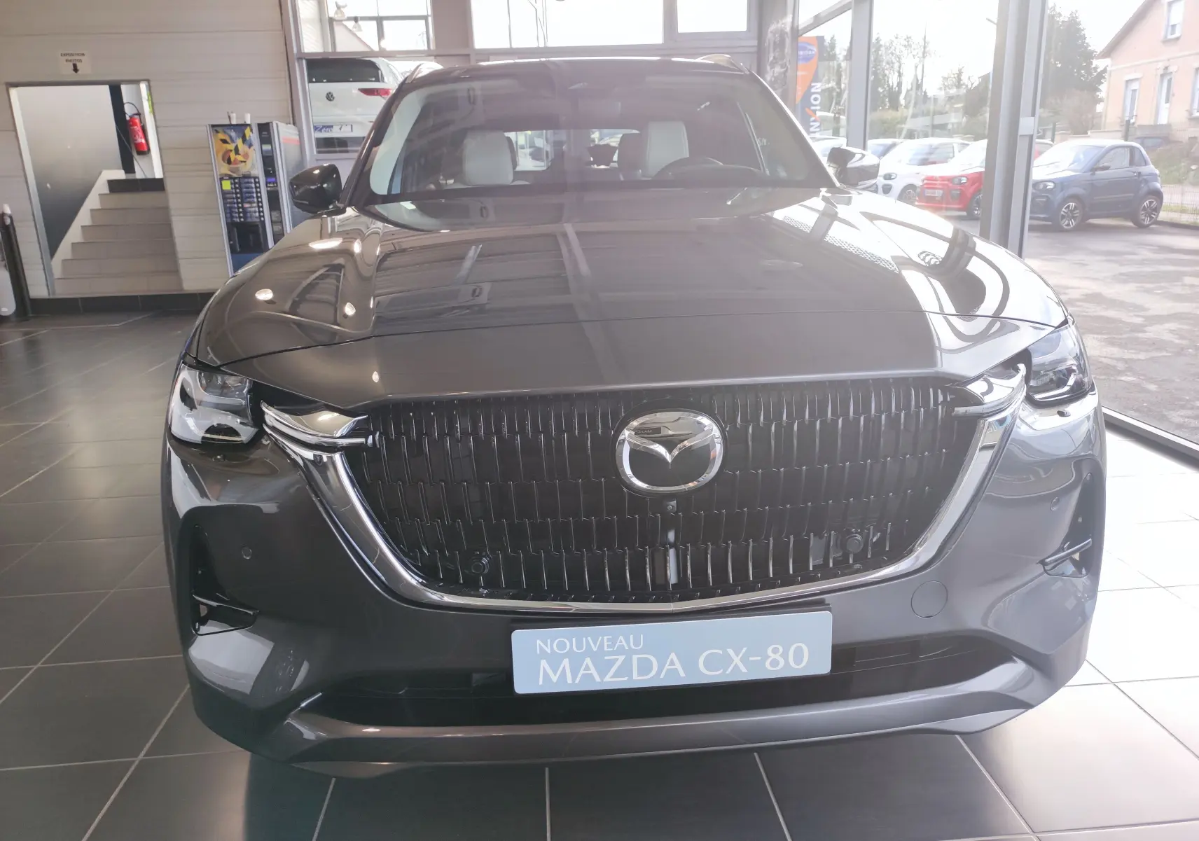 Vue frontale du Mazda CX-80 gris foncé 2024 en showroom, mettant en valeur sa calandre imposante et ses phares LED.