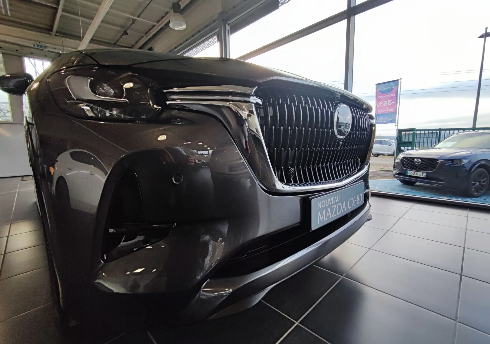 Vue 3/4 avant gauche du Mazda CX-80 gris foncé avec sa calandre imposante et ses phares LED dans un showroom.