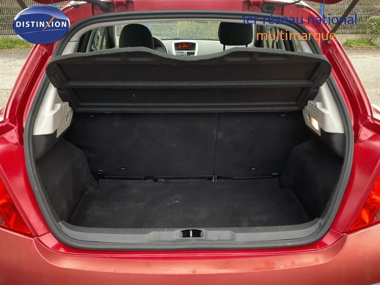 Coffre ouvert d'une Peugeot 207 rouge métal vue arrière, montrant l'espace de chargement et la banquette arrière.