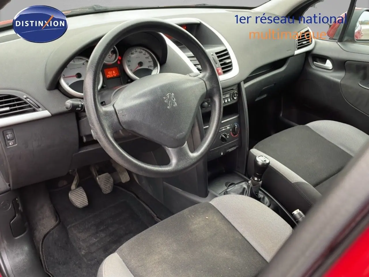 Vue intérieure côté conducteur de la Peugeot 207 rouge métal, montrant le volant, tableau de bord et sièges tissu noir et gris.