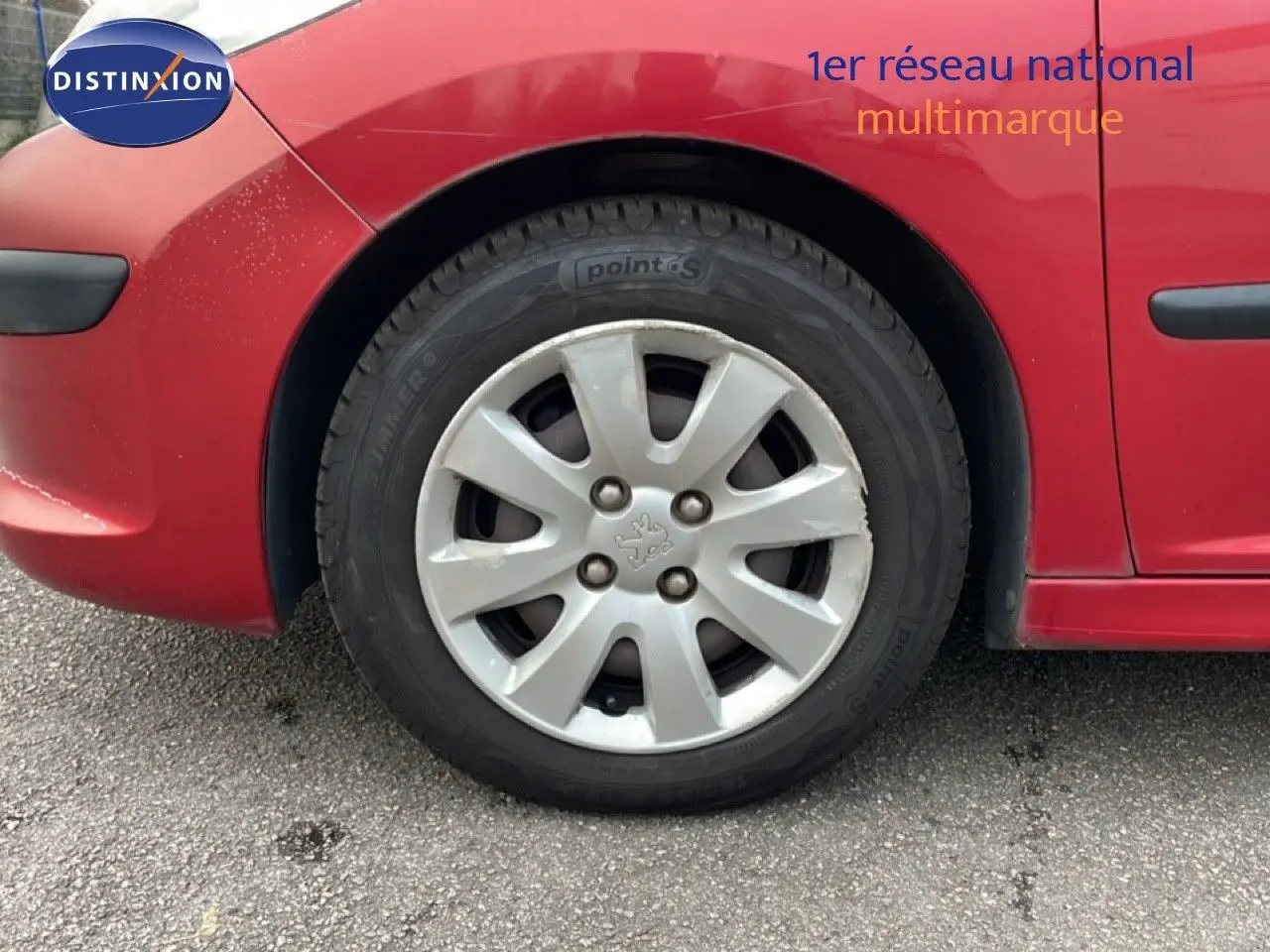 Gros plan sur la roue avant gauche d'une Peugeot 207 rouge métal, mettant en valeur la jante et la carrosserie.