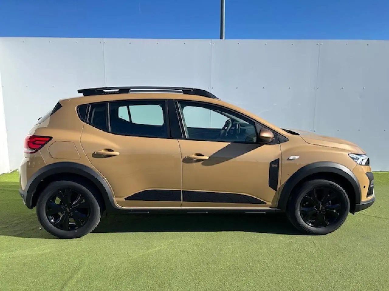 Profil droit d'une Dacia Sandero Stepway beige Safari avec jantes noires et barres de toit noires brillantes.
