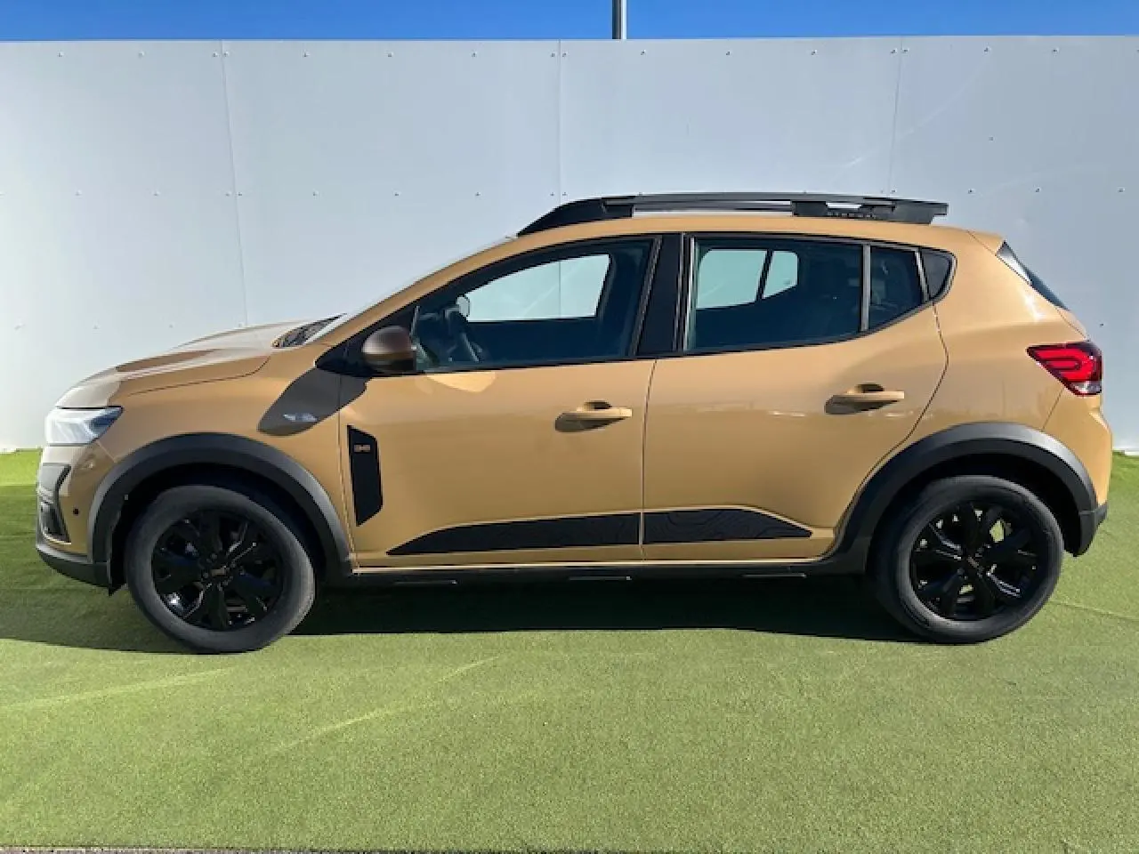 Profil côté gauche de la Dacia Sandero III Stepway beige Safari avec jantes noires et barres de toit gris Mégalithe.