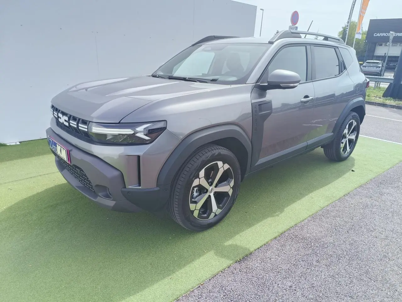Dacia Duster III gris schiste métal en 3/4 avant droit, avec jantes alliage diamantées et barres de toit noires.