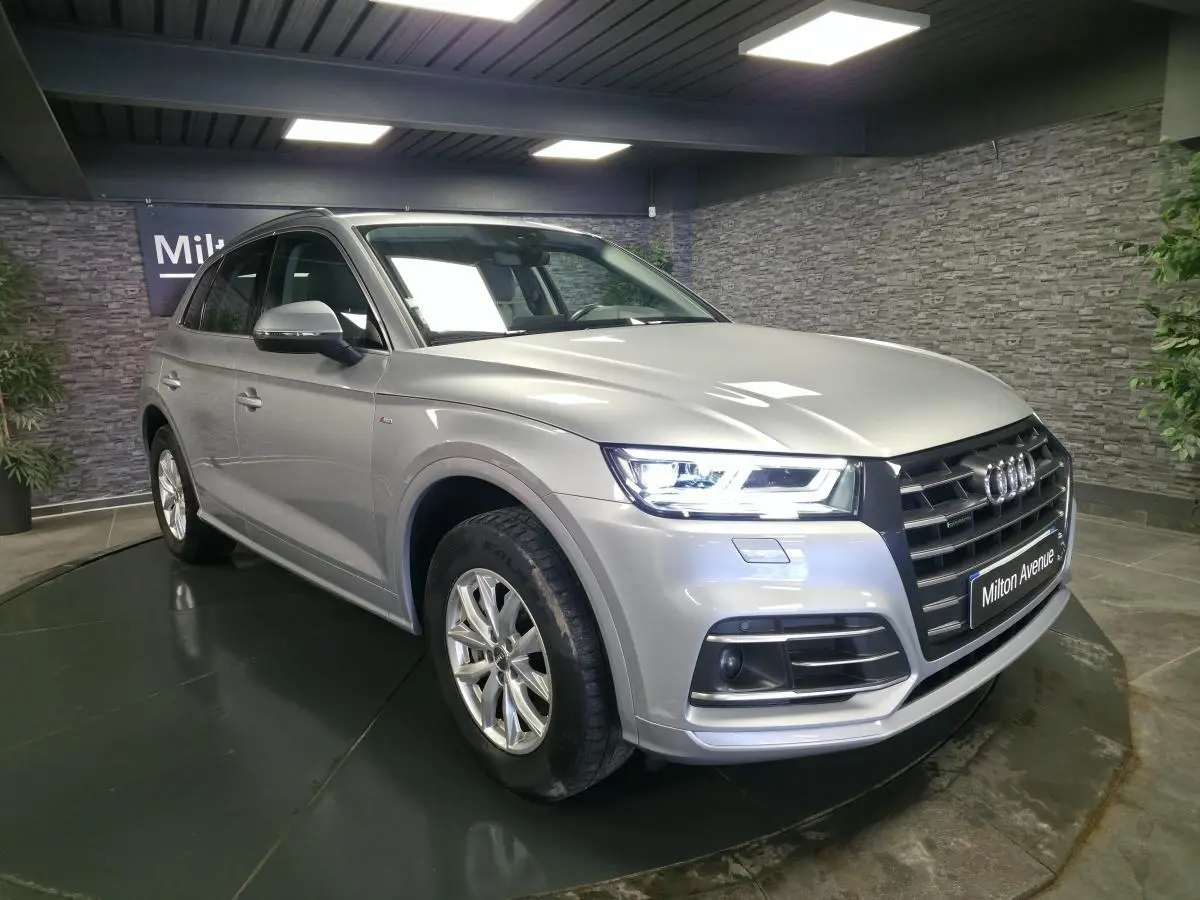 Audi Q5 gris métallisé en 3/4 avant droit, avec calandre noire et phares LED allumés dans un showroom.