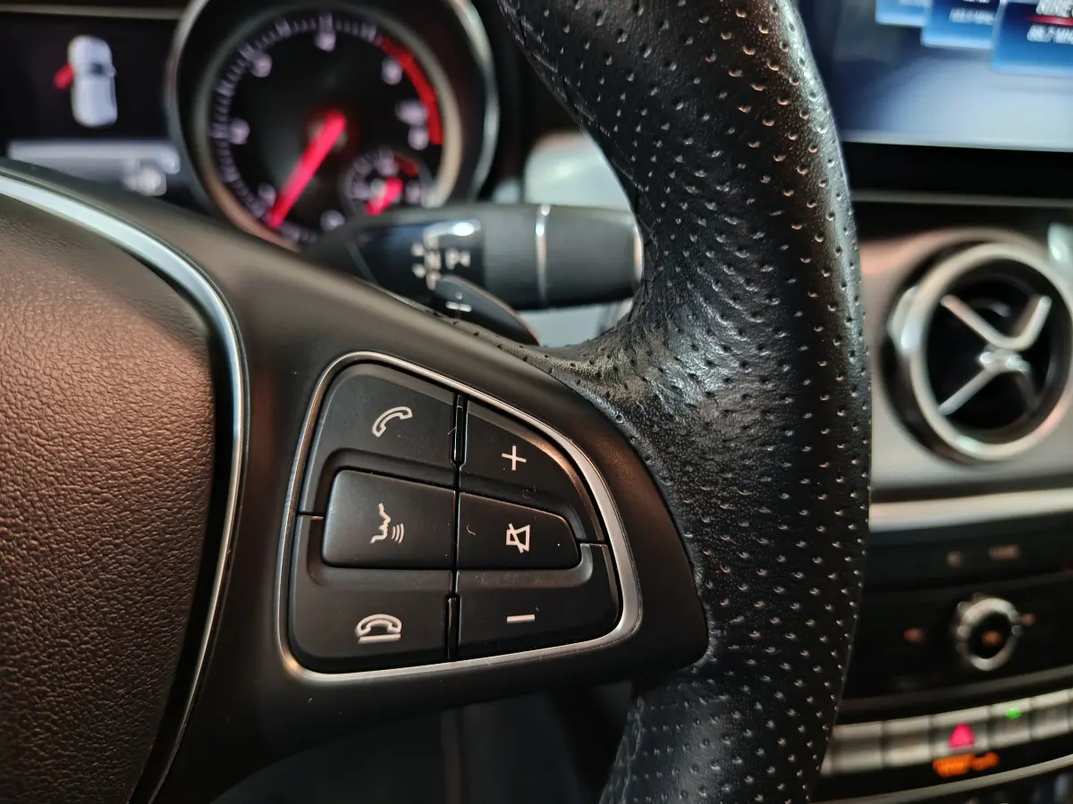 Gros plan sur les commandes gauche du volant cuir perforé du Mercedes GLA 200 d gris clair, avec tableau de bord en arrière-plan.