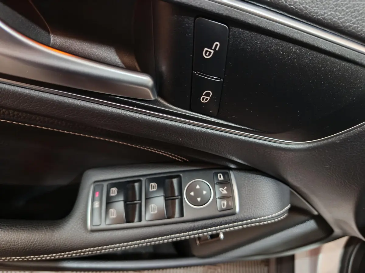 Gros plan sur la commande des vitres et rétroviseurs côté gauche en cuir noir avec surpiqûres blanches dans une Mercedes GLA gris clair.